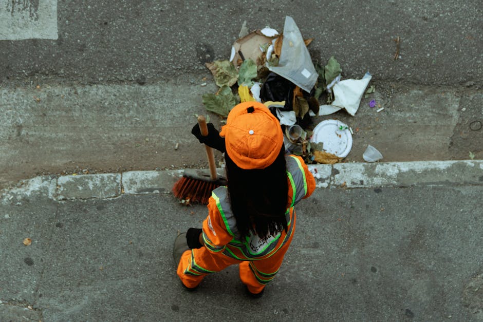 An individual dressed in an orange high-visibility uniform with reflective stripes and an orange cap, viewed from above, is standing on a paved street. The person is holding a small broom in their right hand and is facing a small pile of mixed waste materials that include crumpled paper, leaves, and plastic waste, positioned near the edge of the sidewalk. The waste pile is scattered on the concrete curb and partially onto the asphalt road, which appears weathered with visible cracks and a faded white line marking the road edge. The environment suggests an urban setting, likely part of a rubbish removal or street cleaning activity, and the scene is lit by natural daylight. This image illustrates a scene typical of street waste collection services, such as those provided by private waste handlers like Rubbish Removal Hackney, offering an alternative to council rubbish collections in the area.