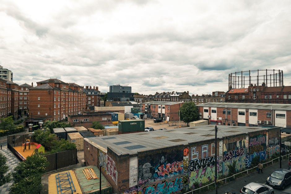 A wide urban scene showing multiple low-rise industrial and residential buildings with brick facades, some with flat roofs covered in metal sheets, and a largeGraffiti-covered wall in the foreground. Several small trees and patches of greenery are visible among the buildings, with a playground featuring a small slide and climbing frame situated on the left side. The background reveals taller brick apartment blocks and a round industrial water tank with a metal railing on top. The sky is overcast with dense, gray clouds, creating a diffused lighting atmosphere typical of cloudy weather. The scene is part of an industrial or storage area, possibly related to waste management or logistics, seen from an elevated perspective, aligning with the context of independent rubbish removal options provided by Rubbish Removal Hackney.