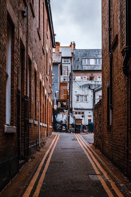 A narrow, paved alleyway flanked by tall red-brick buildings on both sides, with black drainpipes and windows along the walls. The alley has double yellow lines along its edges and appears to be in an urban residential area. At the far end of the alley, there is a small open space featuring a variety of waste and recycling bins, some partially covered with plastic sheetings, indicating private waste collection or disposal arrangements. Behind the bins, a fire escape ladder attached to an adjacent grey building extends upward, and the background shows a mixture of older and modern rooftops, with a cloudy sky overhead. The environment appears damp, suggesting recent rain, and the scene is quiet and utilitarian, consistent with an area where independent waste removal services such as those provided by Rubbish Removal Hackney could operate as an alternative to municipal rubbish collection services.