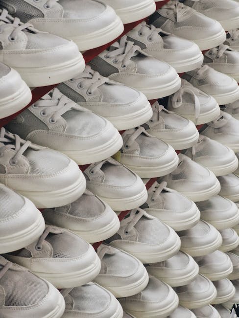 An arranged display of numerous white sneakers with grey accents, neatly stacked on top of each other. The shoes are primarily made of smooth leather and breathable mesh fabric, with white laces tied in standard bows. They are positioned in multiple rows, with some overlapping, and rest on a red surface, possibly a retail shelf or storage display. The environment appears to be indoors, with even lighting that highlights the clean, fresh appearance of the footwear. The organized arrangement of sneakers suggests inventory storage or retail presentation, aligning with aspects of private footwear collection or stock management, which can be associated with alternative waste handling or donation of unused items. This scene emphasizes the volume and uniformity of the shoes, reflecting a focus on bulk storage or disposal of footwear commonly handled through independent collection services like those provided by rubbish removal professionals or dedicated waste management companies.