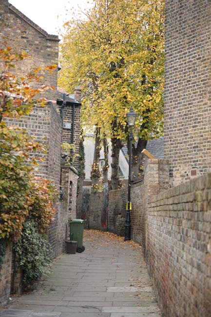A narrow, paved alleyway flanked by tall red-brick buildings on both sides, with black drainpipes and windows along the walls. The alley has double yellow lines along its edges and appears to be in an urban residential area. At the far end of the alley, there is a small open space featuring a variety of waste and recycling bins, some partially covered with plastic sheetings, indicating private waste collection or disposal arrangements. Behind the bins, a fire escape ladder attached to an adjacent grey building extends upward, and the background shows a mixture of older and modern rooftops, with a cloudy sky overhead. The environment appears damp, suggesting recent rain, and the scene is quiet and utilitarian, consistent with an area where independent waste removal services such as those provided by Rubbish Removal Hackney could operate as an alternative to municipal rubbish collection services.