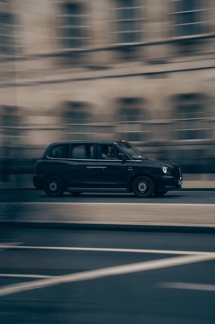 A black London taxi cab is captured in motion driving along a city street with a blurred background of multi-storey buildings, indicating rapid movement. The vehicle’s exterior features a glossy finish with visible windows, door handles, and the iconic taxi shape. The pavement and road markings are visible beneath the cab, with subtle reflections on the wet surface suggesting recent rain. The scene is illuminated by natural daylight, creating a neutral, professional atmosphere appropriate for urban transport contexts. This image could subtly relate to alternative transportation methods often contrasted with private waste disposal or rubbish collection services, emphasizing the variety of urban mobility and logistics options. Overall, the scene highlights the movement of an established taxi service within a densely built city environment, reflecting the fast pace of urban life and the importance of efficient vehicle use in metropolitan areas, which can connect indirectly to themes of waste collection and service logistics managed by companies like Rubbish Removal Hackney in the context of alternative rubbish handling solutions.