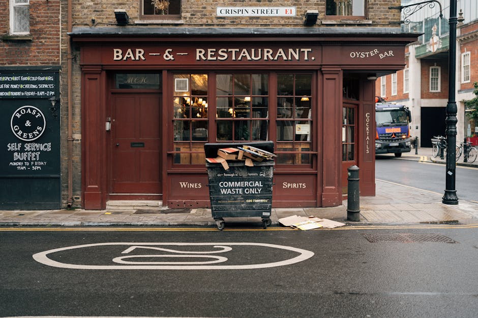 On a city street in front of a reddish-brown bar and restaurant with large glass windows and wooden framing, a black commercial waste bin with white lettering indicating it is for waste disposal only stands on the pavement. The bin is overflowing with flattened cardboard boxes and paper waste, some of which have spilled onto the ground nearby. To the right of the bin, a short black bollard is positioned at the edge of the sidewalk, separating the pedestrian area from the road. The asphalt road features a white no-parking symbol painted prominently in the foreground. In the background, a blue delivery truck is parked further along the street, and there are a few bicycles leaning against a lamppost. The scene is illuminated by natural daylight with a slightly overcast sky, providing a neutral and clear view of the urban environment, reflecting the backdrop of a typical location where private rubbish removal and waste collection services might be employed as an alternative to local authority bin collection.