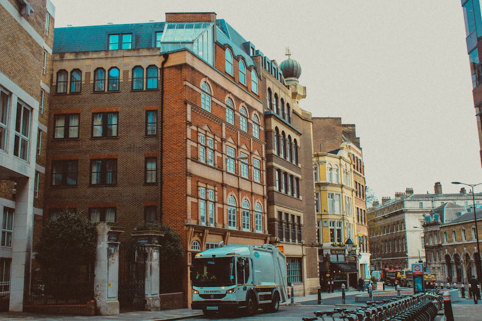 A street scene in an urban area featuring a row of historic multi-storey buildings with brick and stone facades, large arched windows, and decorative architectural details. The buildings vary in color, including warm tones of red brick, cream, and pale yellow, with some featuring ornate cornices and bay windows. In the foreground, a rubbish collection vehicle painted in light blue is parked on the pavement near a gated entrance with stone pillars, indicating an example of private waste collection or alternative rubbish removal methods. Bicycles are lined up along a bike rack on the right side of the image, while a street lamp and a few pedestrians are visible in the background, suggesting a busy city centre setting. The overall scene is captured in natural daylight with overcast skies, emphasizing the architectural details and urban environment suitable for a rubbish removal service overview in a historical district.