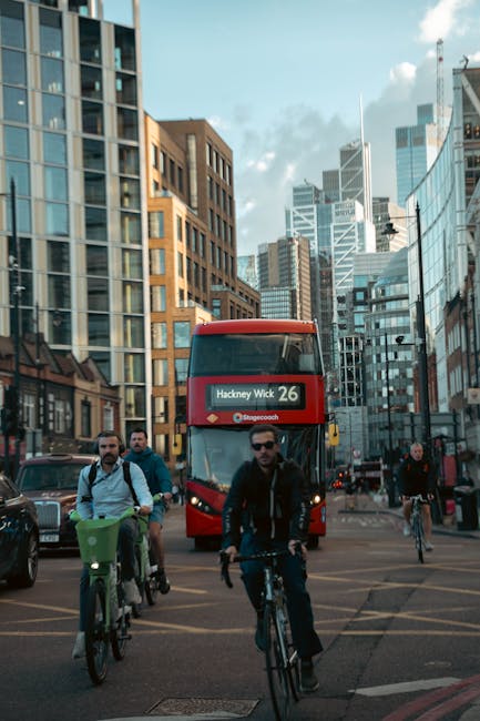 A busy urban street scene in Hackney, showing several cyclists riding across a wide intersection in the foreground, with a young man wearing sunglasses, a dark jacket, and held handlebars on his bike. To his left, there is a person on a green bicycle with a basket attached to the front. In the background, a red double-decker bus displaying the route number 26 and the destination 'Hackney Wick' is positioned amidst modern high-rise buildings with glass and brick facades, reflecting the daylight and sky above. The scene is illuminated by natural daylight, with some clouds visible in the sky, and the surrounding environment includes traffic lights, road markings, and a few parked cars, indicating an active city street front typical of Hackney. The overall atmosphere captures a vibrant, active city environment with a focus on urban transportation and active commuting, relevant to services like private rubbish and waste collection that support maintaining the cleanliness of such busy city areas.